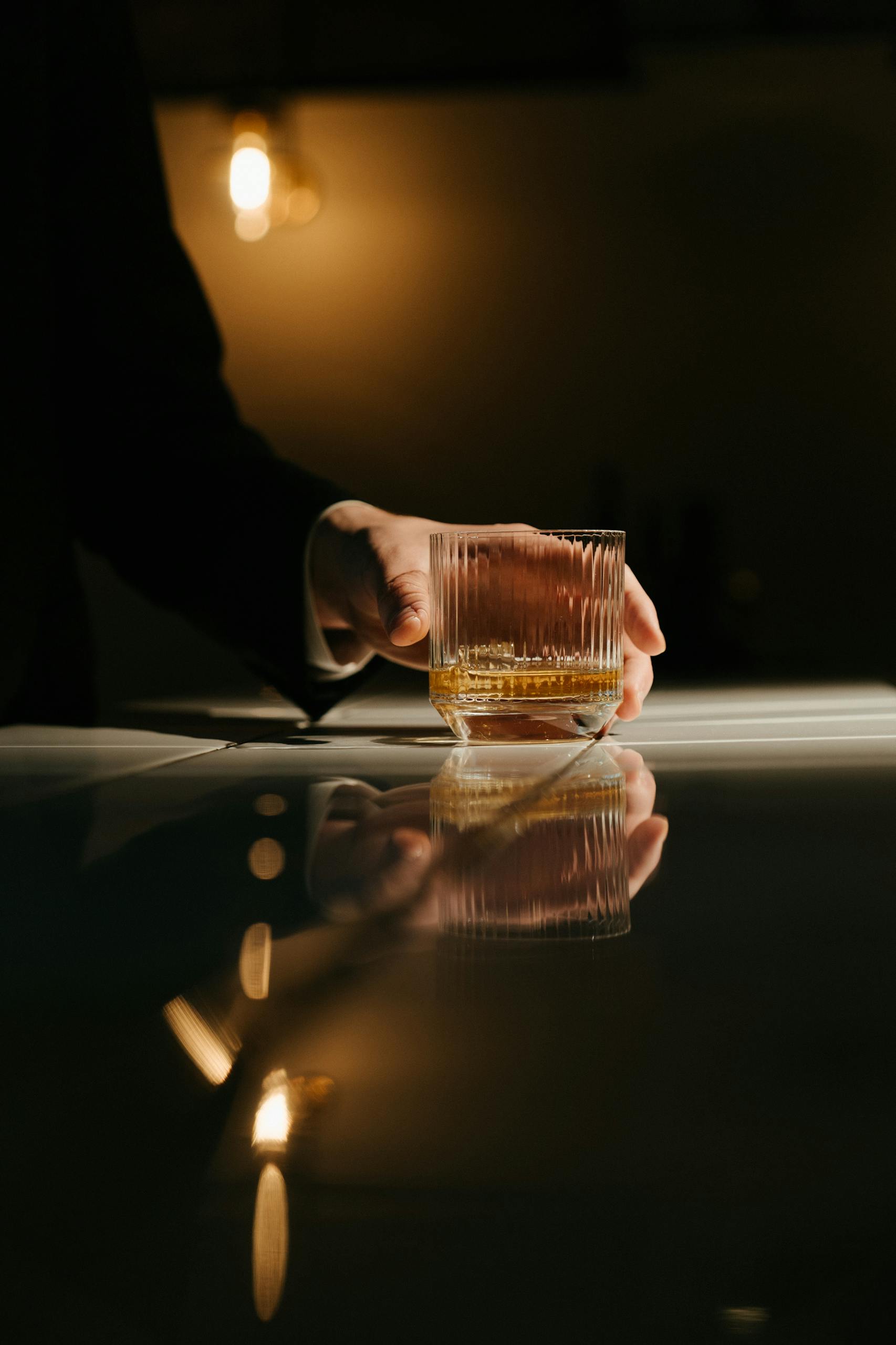 Close-up of a hand holding a whiskey glass with reflections on a bar counter in a dim light. What we treat