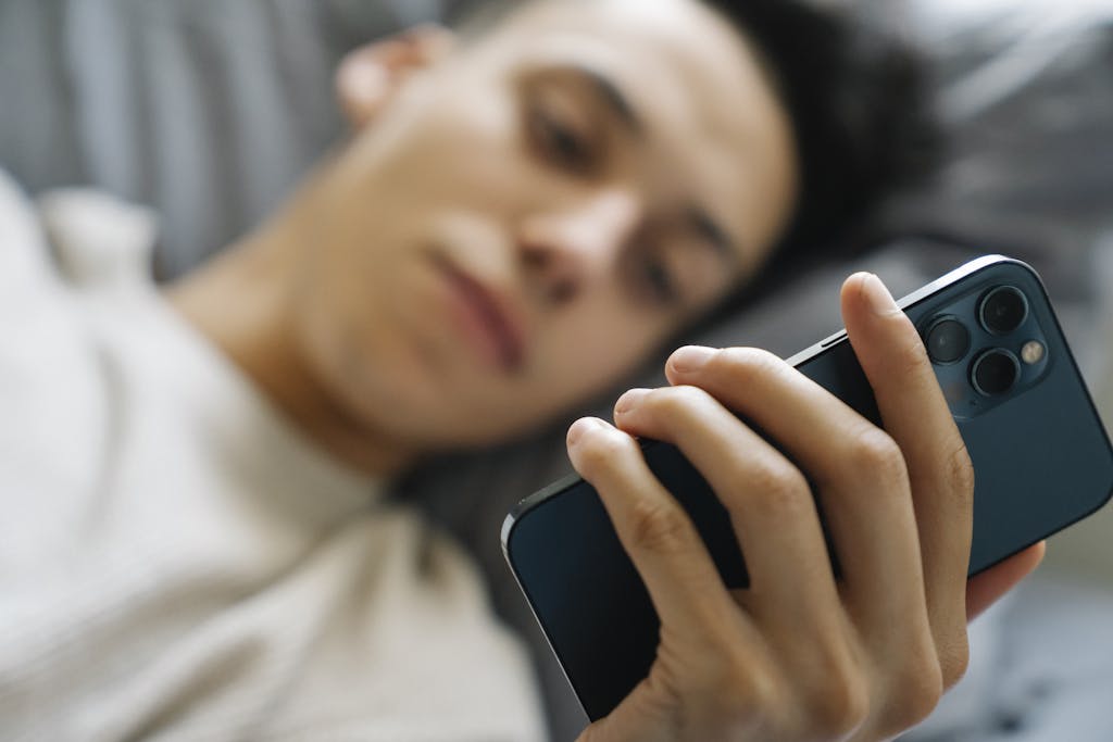 Soft focus of young guy lying on bed and using modern smartphone in weekend morning at home